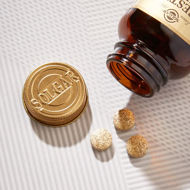A Solgar Digestive Enzymes 100 Tablets bottle, brown with a gold cap off, lies on its side with three round tablets next to it on a textured white surface.