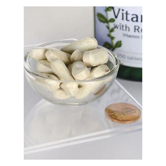 A small glass bowl with beige tablets sits on a clear surface beside a coin for scale. In the background, a partially visible bottle labeled "Vitamin C-1000 with Rose Hips 250 Tablets" by Swanson hints at its powerful antioxidants.