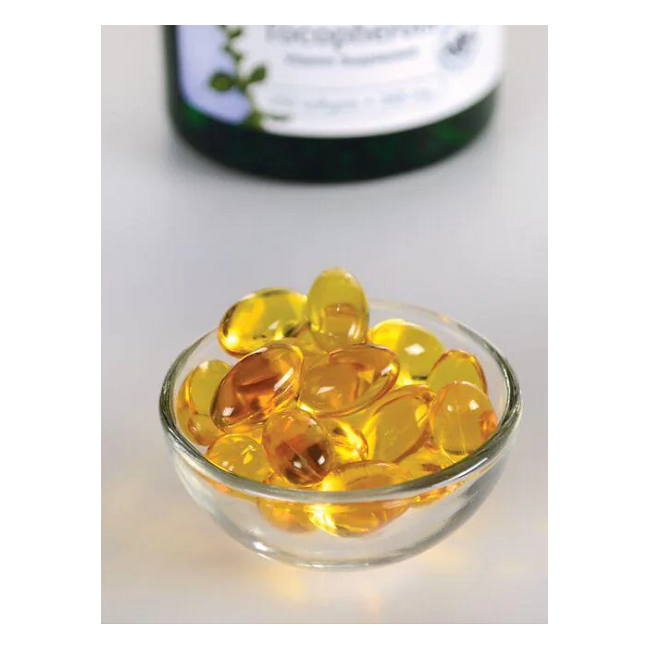 A small glass bowl with Swanson's Vitamin E-400 Mixed Tocopherols softgels, rich in antioxidants, sits on a white surface. A blurred bottle is visible in the background.