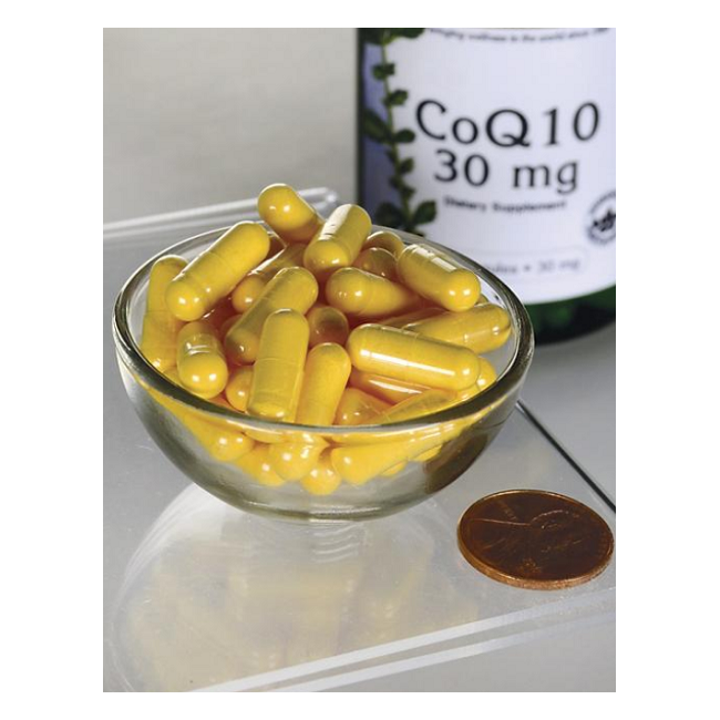 A glass bowl of small yellow capsule pills is placed next to a penny for size reference. In the background, a Swanson "Coenzyme Q10 30 mg 240 Capsules" bottle is visible, emphasizing its role in energy production and supporting cardiovascular health.