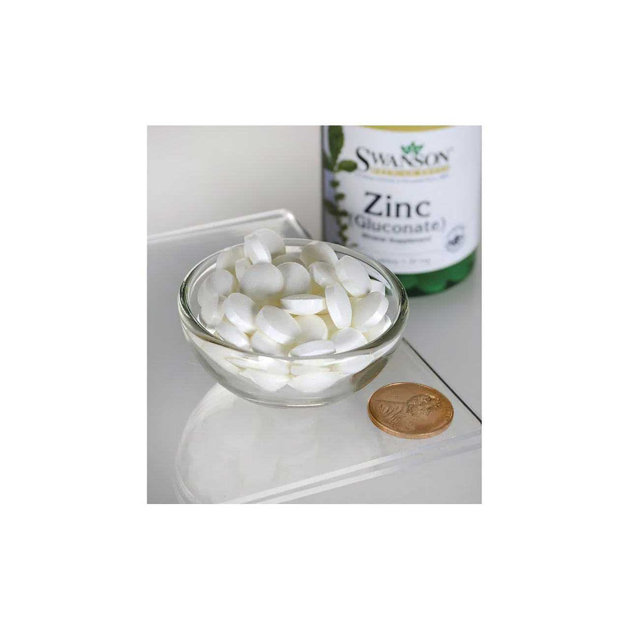 A bowl of white capsules next to a bottle of Swanson Zinc Gluconate 30 mg 250 Tablets supplements for daily wellness.