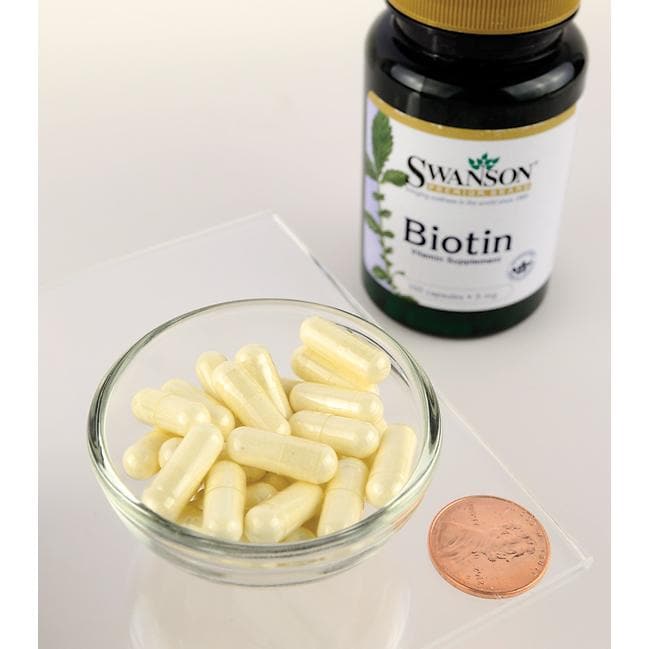 A dietary supplement bottle of Swanson Biotin - 5 mg 100 capsules next to a penny on a table.